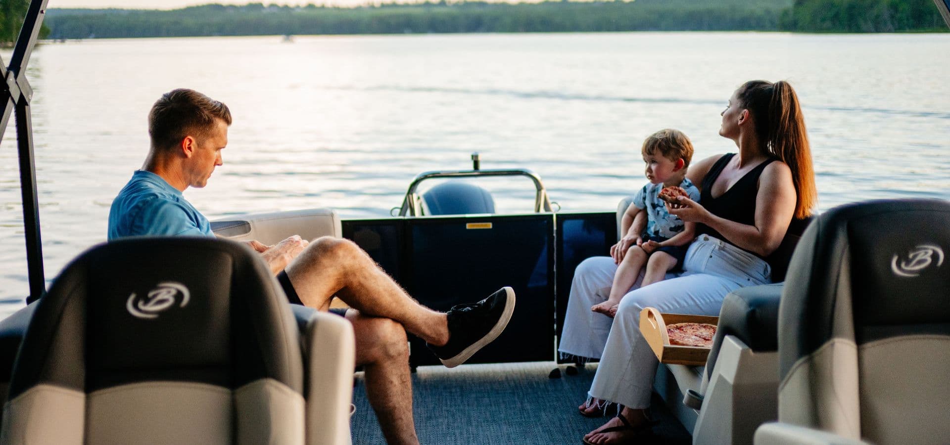 A man sits on a boat while a woman holds a child and enjoys the lakeside view.