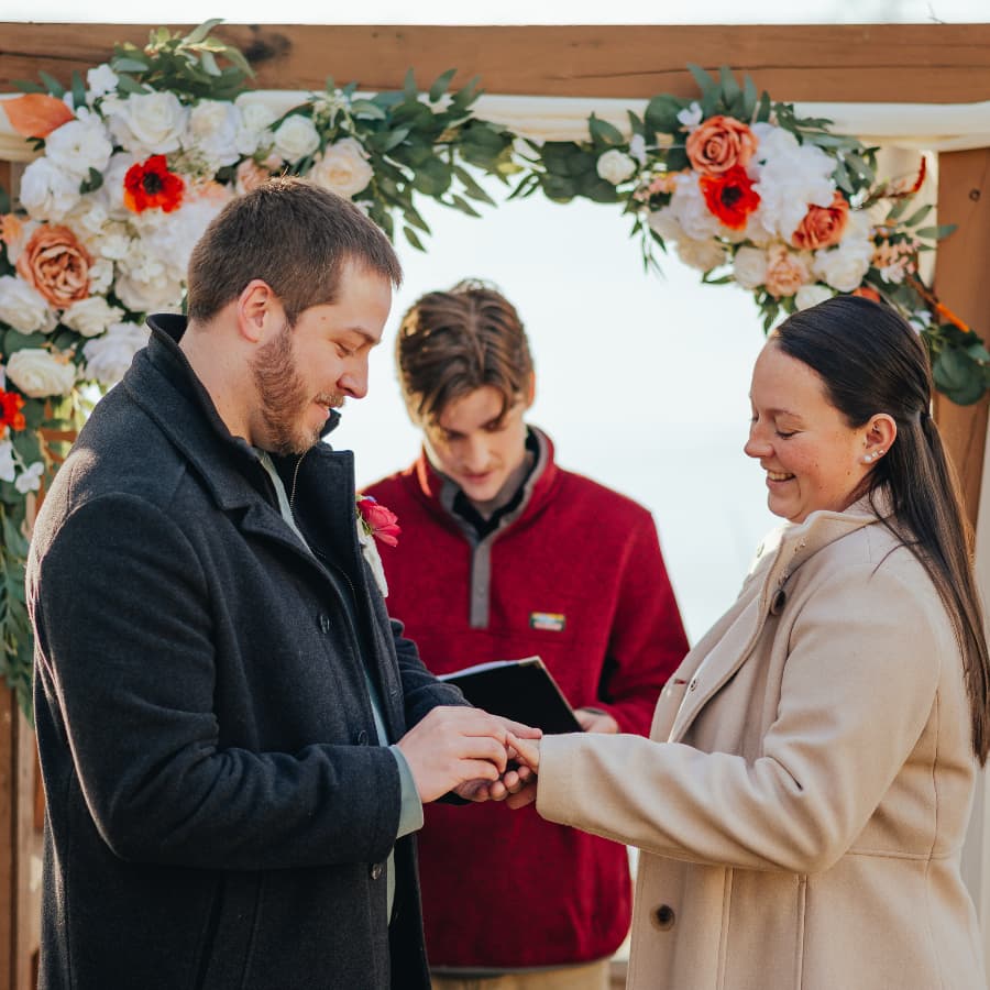 A couple exchanges rings during their wedding ceremony under a floral arch.
