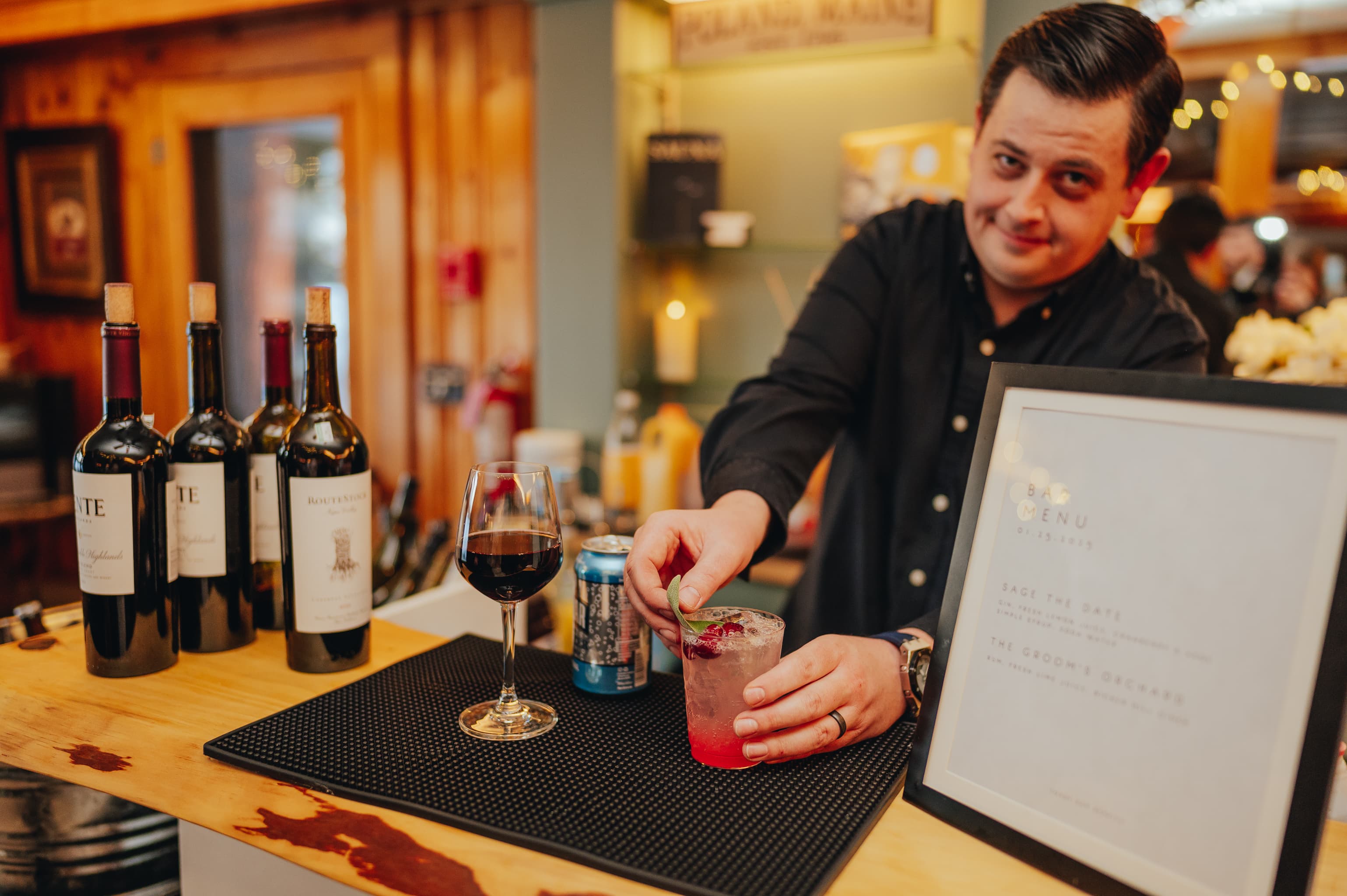 A bartender mixes a colorful cocktail while red wine bottles and a menu are visible on the bar.