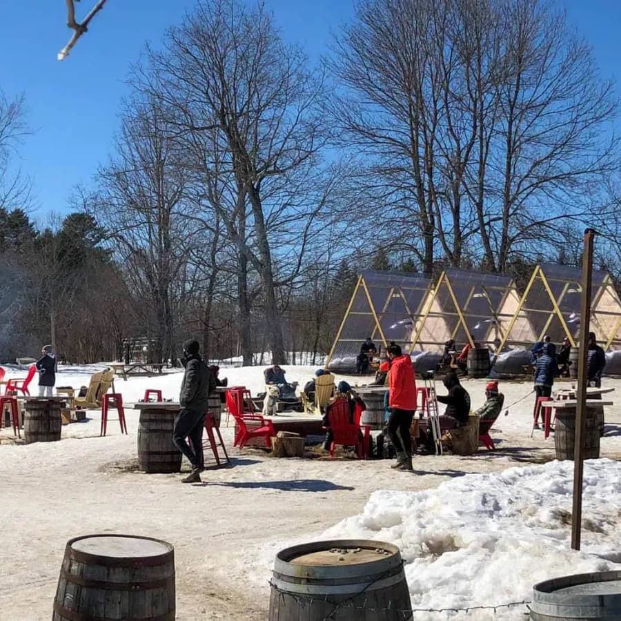 A snowy outdoor area with people seated in red chairs around fire pits and wooden barrels, surrounded by trees and structures.