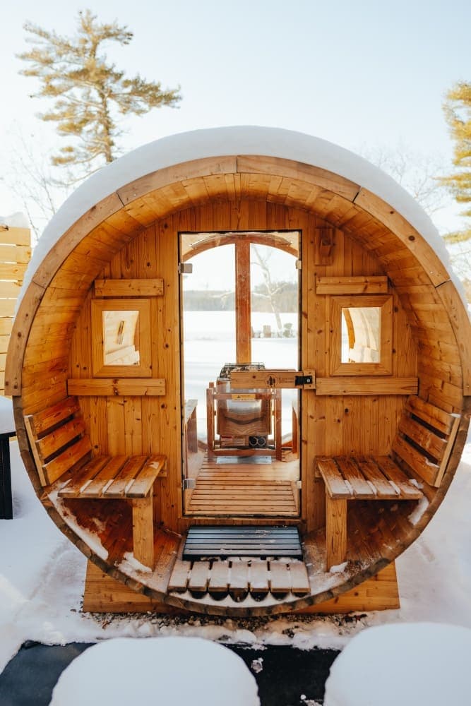 A round wooden sauna with benches, partially covered in snow, set against a winter landscape.
