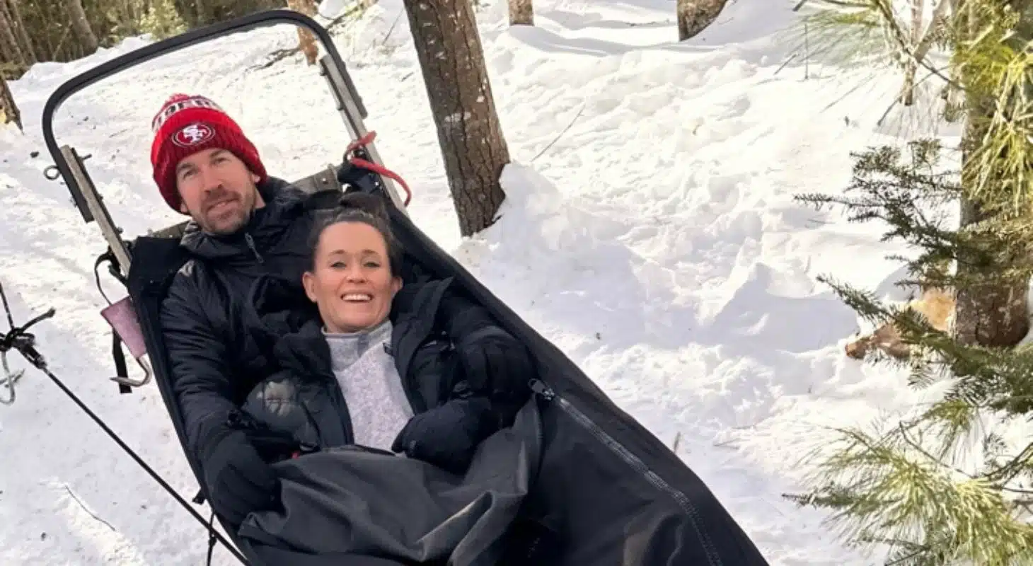 A man and woman smile while sitting together in a snow-covered landscape, wrapped in blankets.