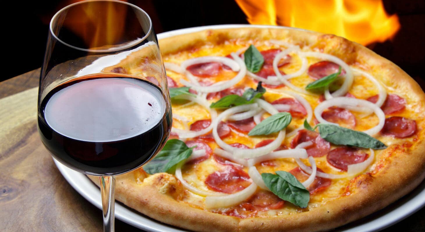A glass of red wine beside a pepperoni and onion pizza garnished with basil, with flames in the background.
