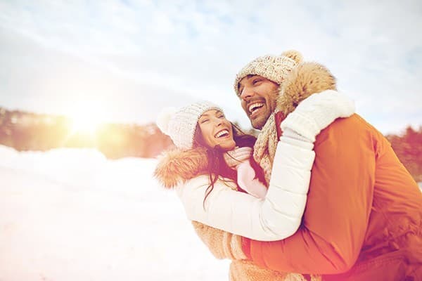 A joyful couple embraces in a snowy landscape, smiling warmly at each other as sunlight breaks through the clouds.