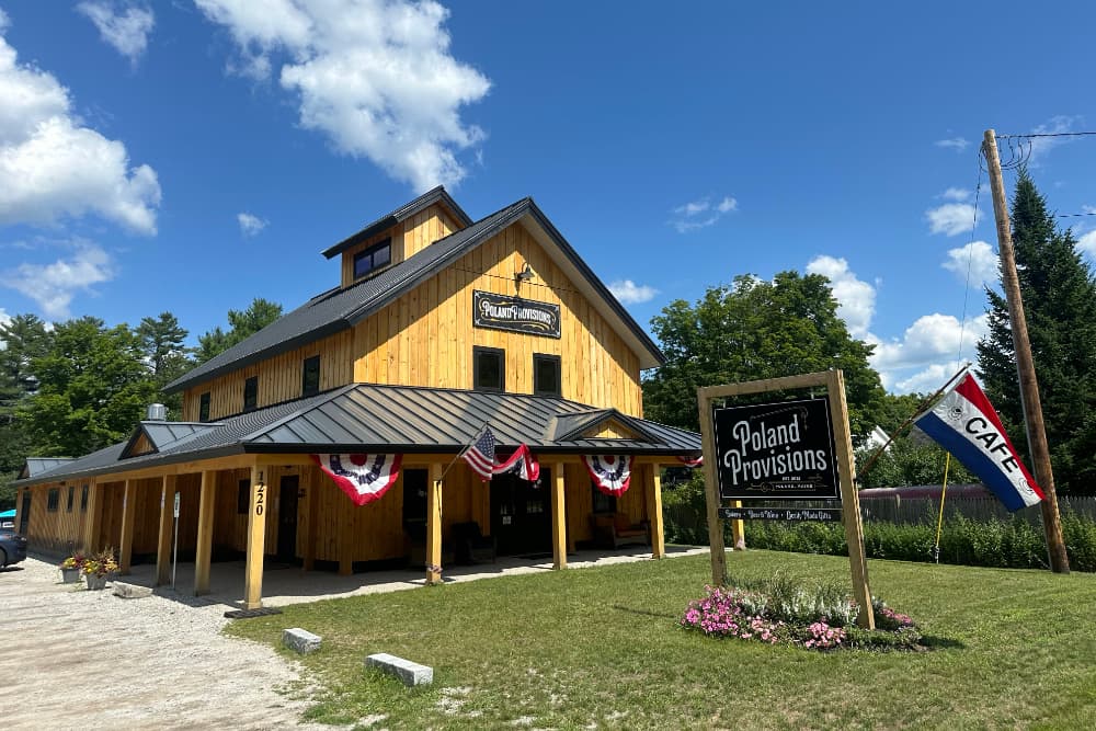 A wooden building with a black roof and "Poland Provisions" sign, featuring red, white, and blue decorations and a café sign, set against a sunny blue sky.