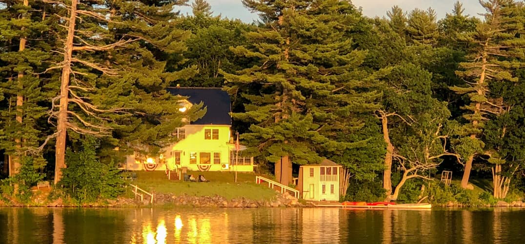 A serene lakeside view featuring a yellow house surrounded by tall trees at sunset.