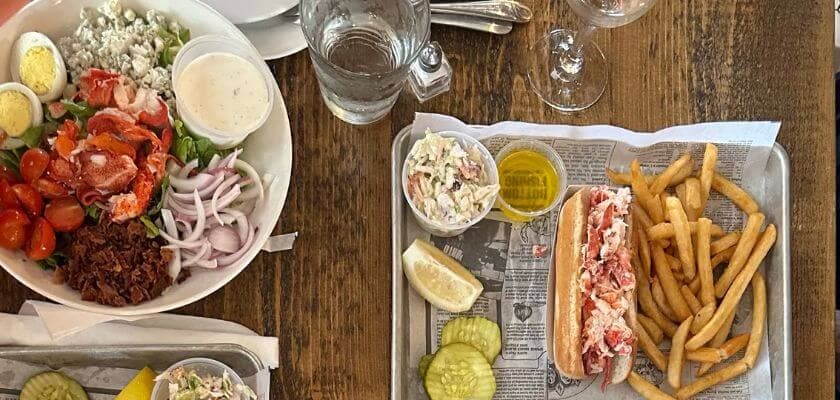 A seaside meal featuring a lobster roll, fries, a green salad, and condiments on a wooden table.