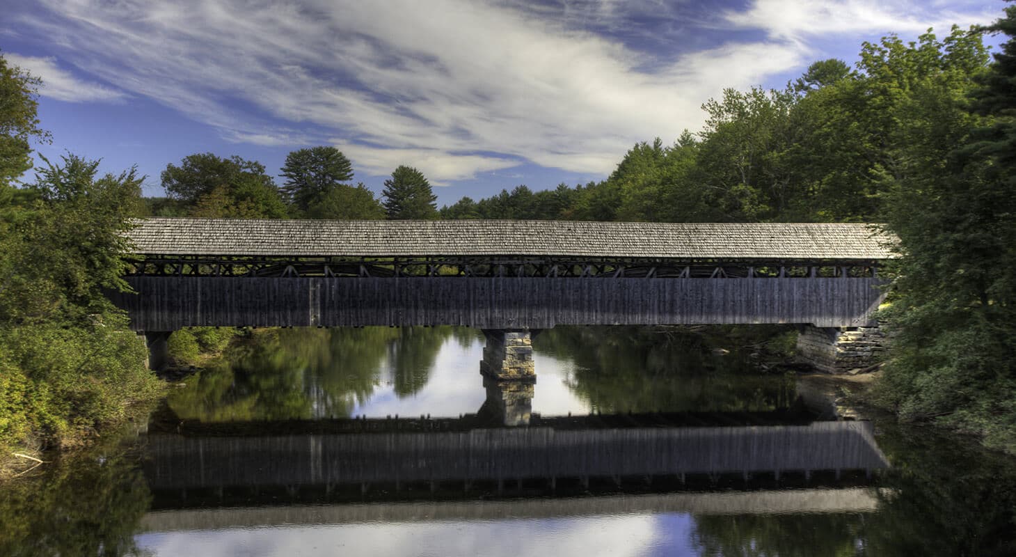 A covered wooden bridge reflects in calm waters under a blue sky with scattered clouds.