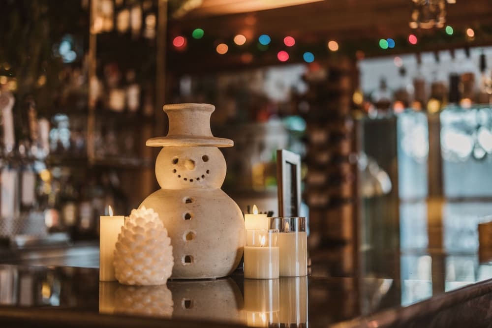 A ceramic snowman figurine is surrounded by candles on a countertop, with festive lights in the background.
