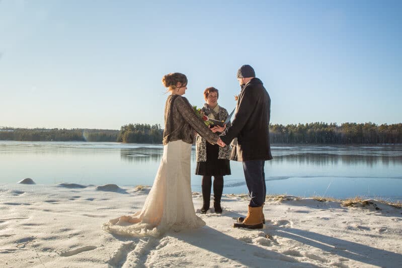 A bride and groom hold hands during their winter wedding ceremony by a frozen lake, with an officiant nearby.