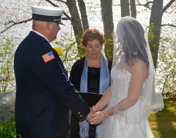 A couple stands hand in hand during their wedding ceremony by a serene lake, with an officiant in the background.