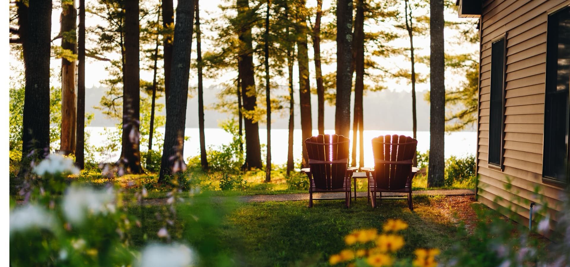 Two red Adirondack chairs sit by a lake, surrounded by trees and flowers at sunset.