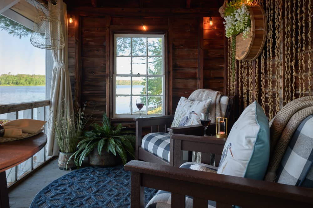 Cozy interior of a lakeside cabin with two armchairs, decorative pillows, wine glasses, and a view of the water through a large window.