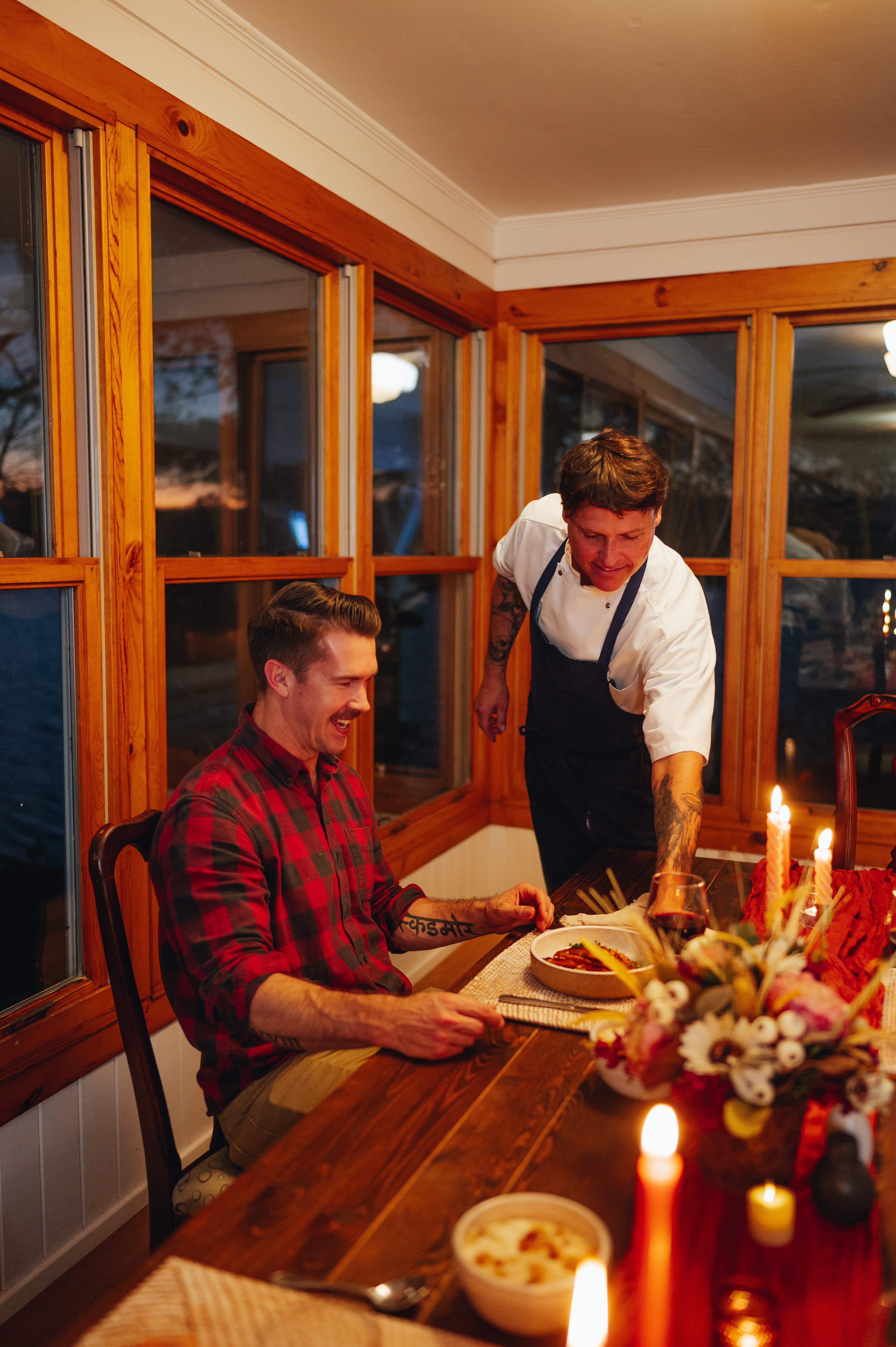 A smiling man in a plaid shirt enjoys a dinner served by a chef in an apron at a well-lit table.