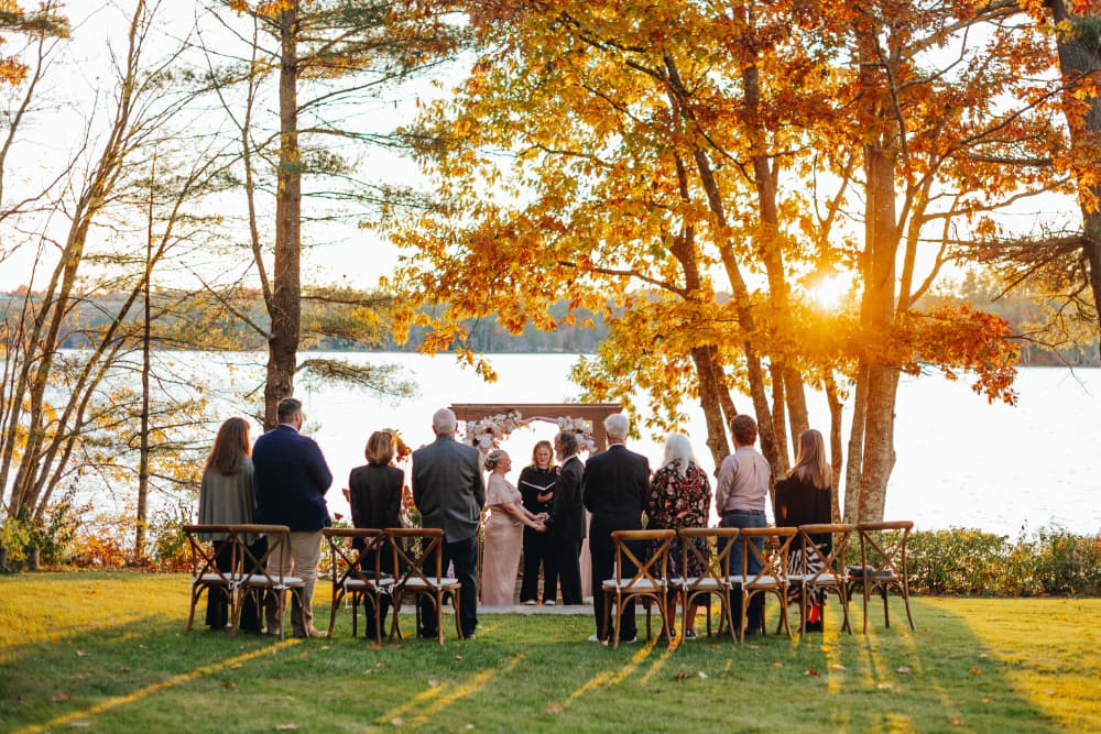 A wedding ceremony takes place by a lake surrounded by autumn trees at sunset.