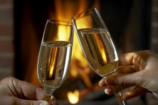 Two champagne flutes clink together in front of a warm fire.
