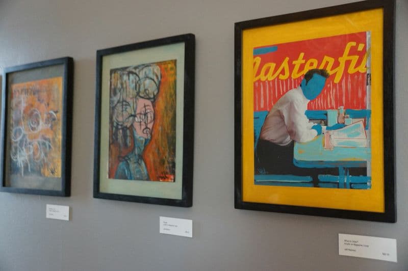 Three framed paintings displayed on a gray wall, featuring abstract art and a figure at a table.