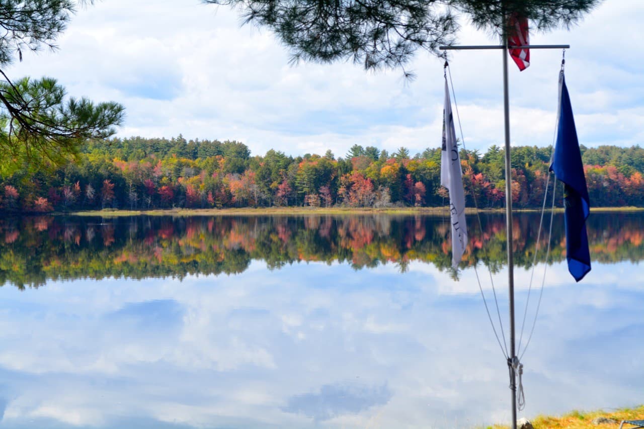 A serene lake reflecting colorful autumn foliage under a cloudy sky, with flags in the foreground.