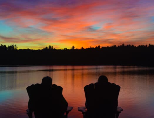 Two silhouettes sit in chairs by a lake, gazing at a vibrant sunset.