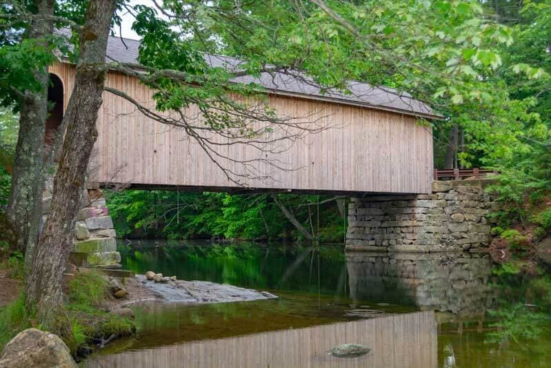 A wooden covered bridge spans a calm river, surrounded by lush green foliage.