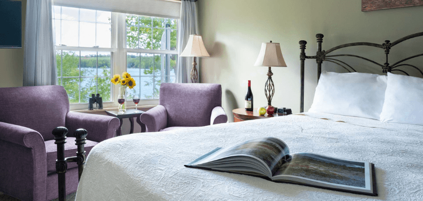 A cozy bedroom featuring a large bed, two purple armchairs, a window with a view of the water, and a book on the bed.