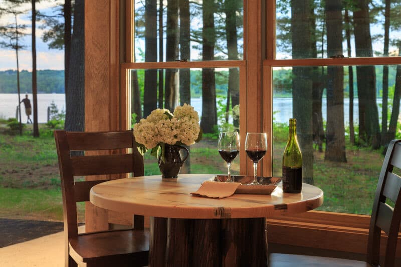 A cozy dining table with two glasses of red wine, a bottle, and flowers, overlooking a serene lake through large windows.