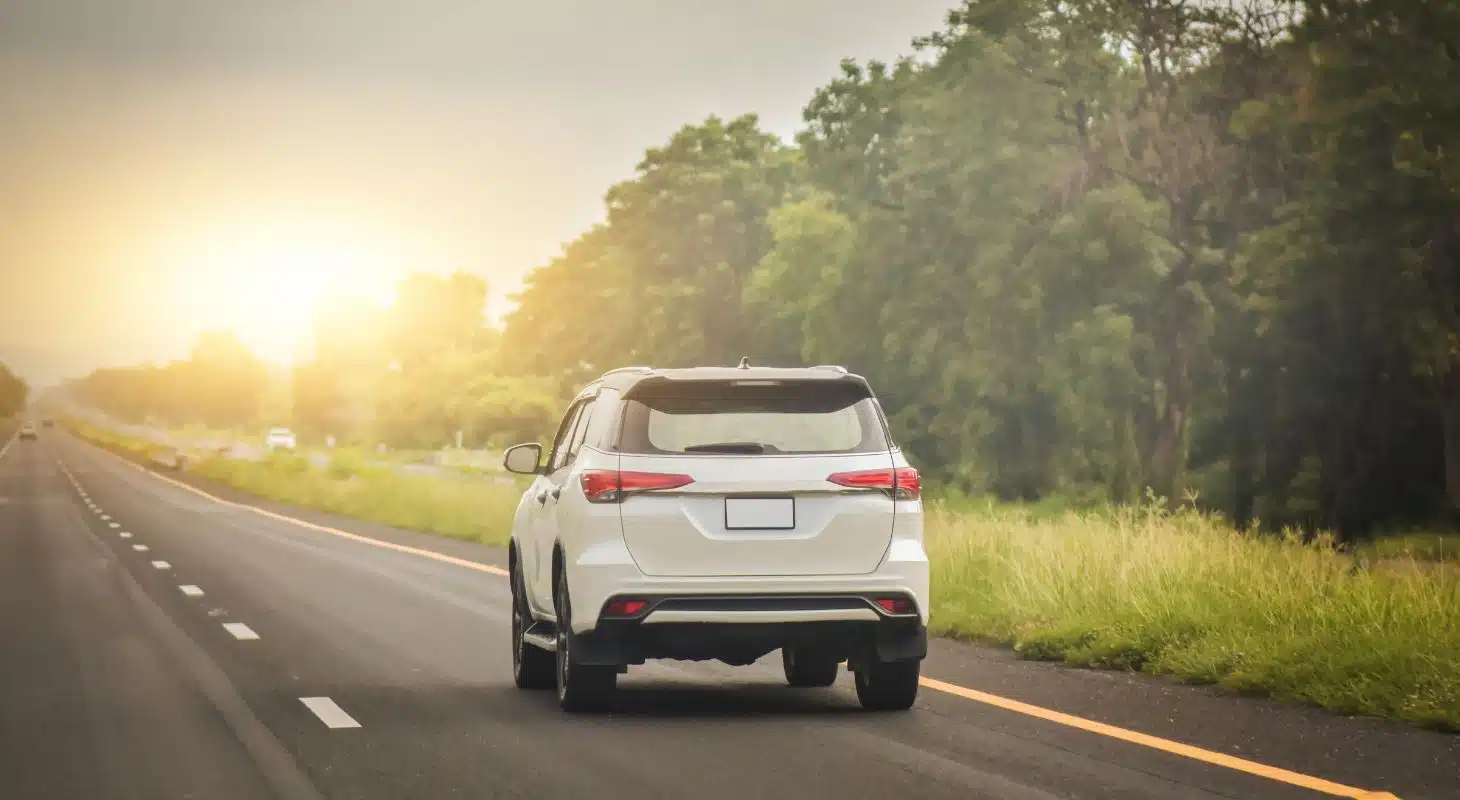 A white SUV driving on a road surrounded by greenery at sunset.