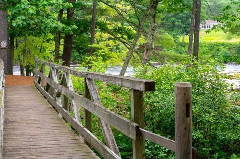 A wooden bridge leads through lush greenery beside a river.