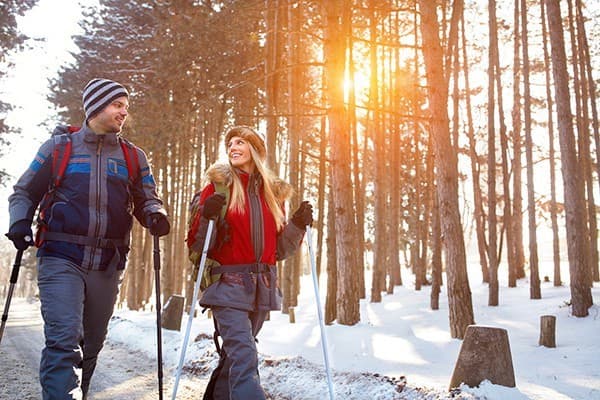 A man and woman hike together through a snowy forest, smiling as the sun sets behind the trees.