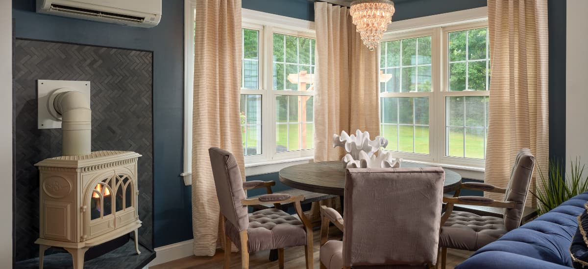A cozy dining area featuring a round table, plush chairs, a decorative stove, and large windows with striped curtains.