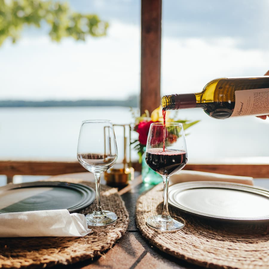 A hand pours red wine into a glass at a beautifully set table by a window overlooking water.
