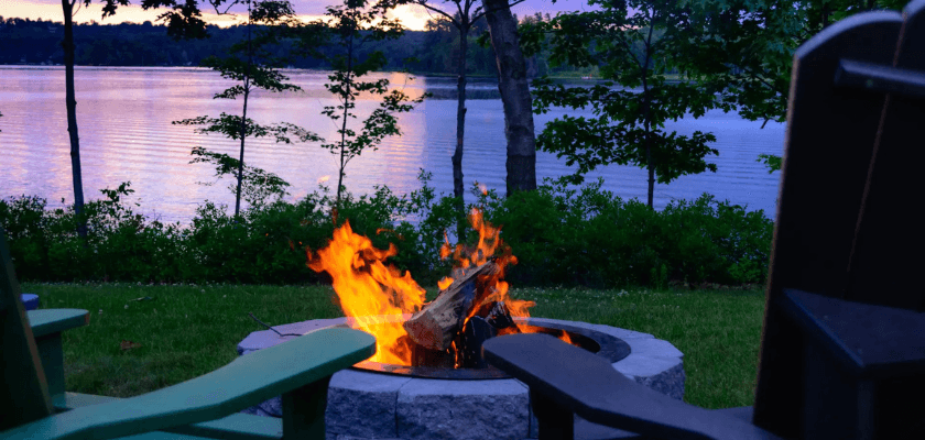 A cozy campfire burns in a stone pit by a serene lakeside at dusk.