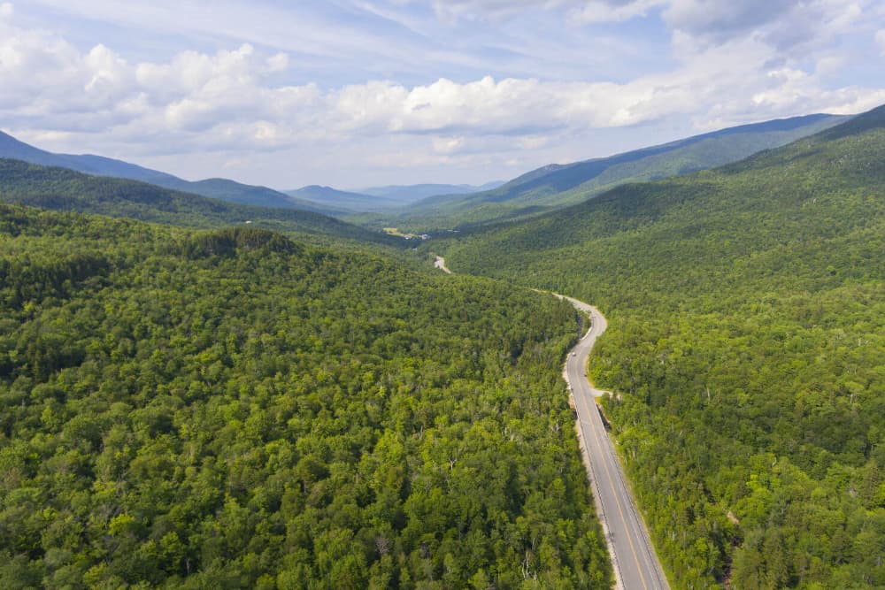 Aerial view of a winding road through lush green mountains and forests under a cloudy sky.
