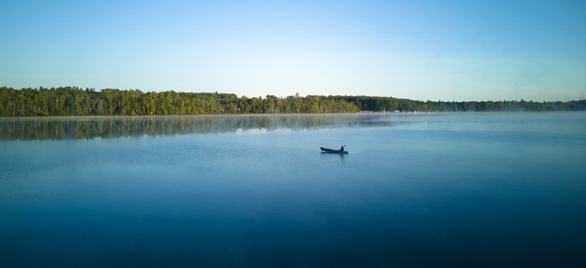 A lone boat floats on a calm lake surrounded by misty trees under a clear blue sky.