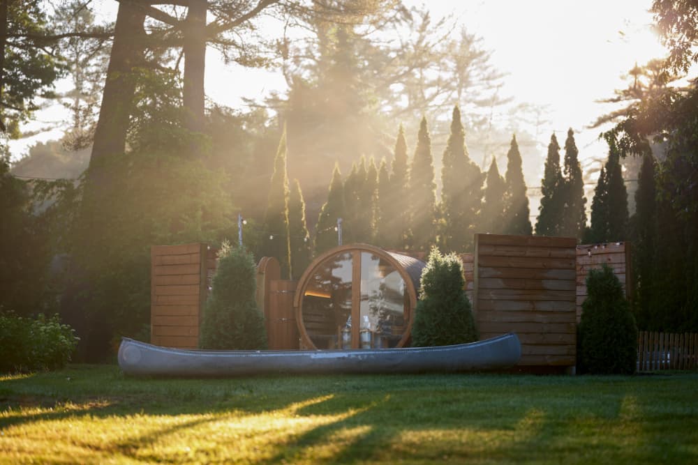 A canoe rests in a sunlit garden featuring a wooden sauna and evergreen trees.