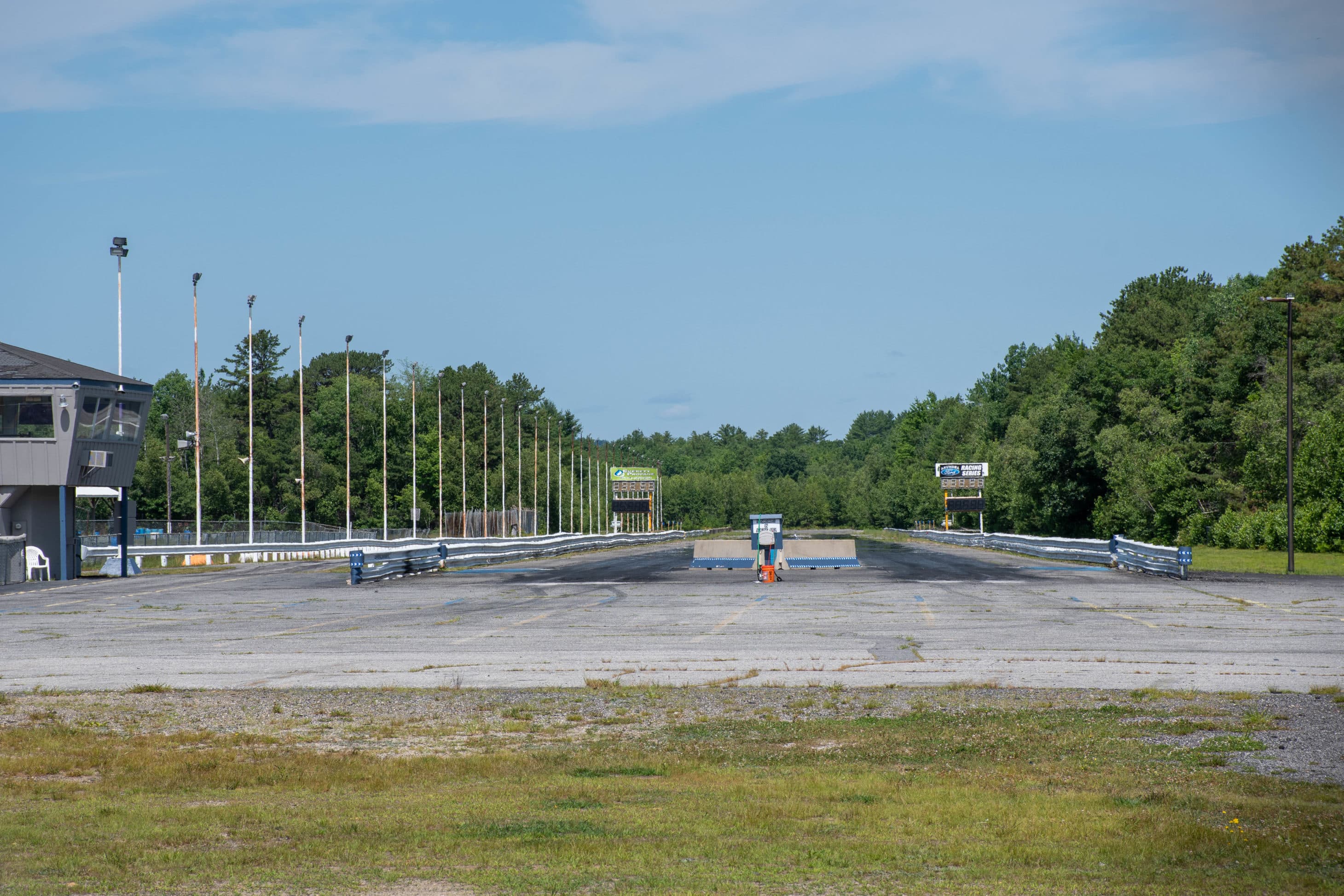 An empty drag racing track lined with light poles and surrounded by trees under a clear blue sky.