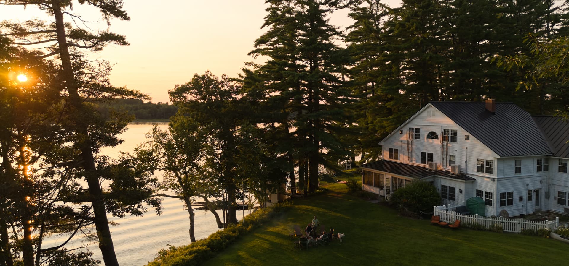 A scenic lakefront view at sunset featuring a large white house surrounded by trees and a gathering of people.
