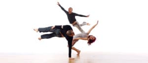 Three dancers performing dynamic movements on a wooden floor against a white backdrop.