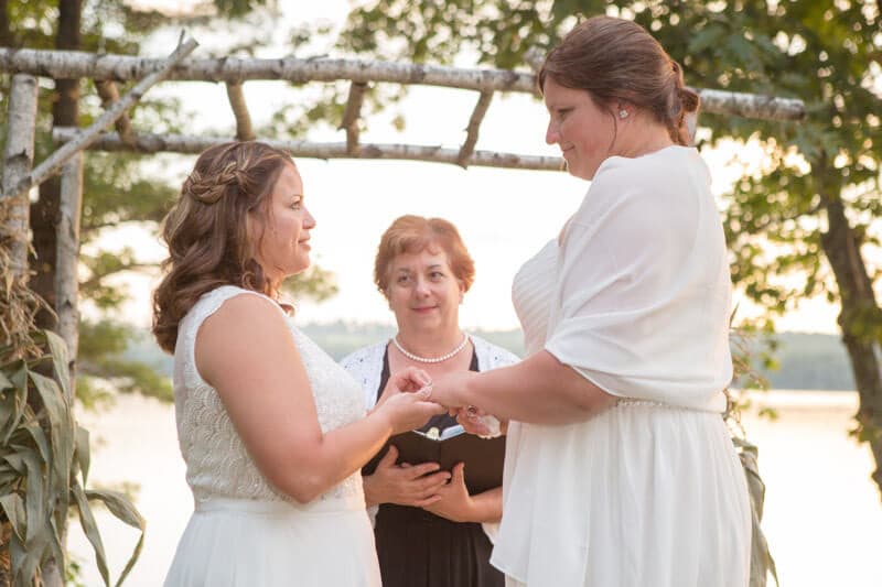 Two women exchange wedding rings while a woman officiates in a serene outdoor setting.