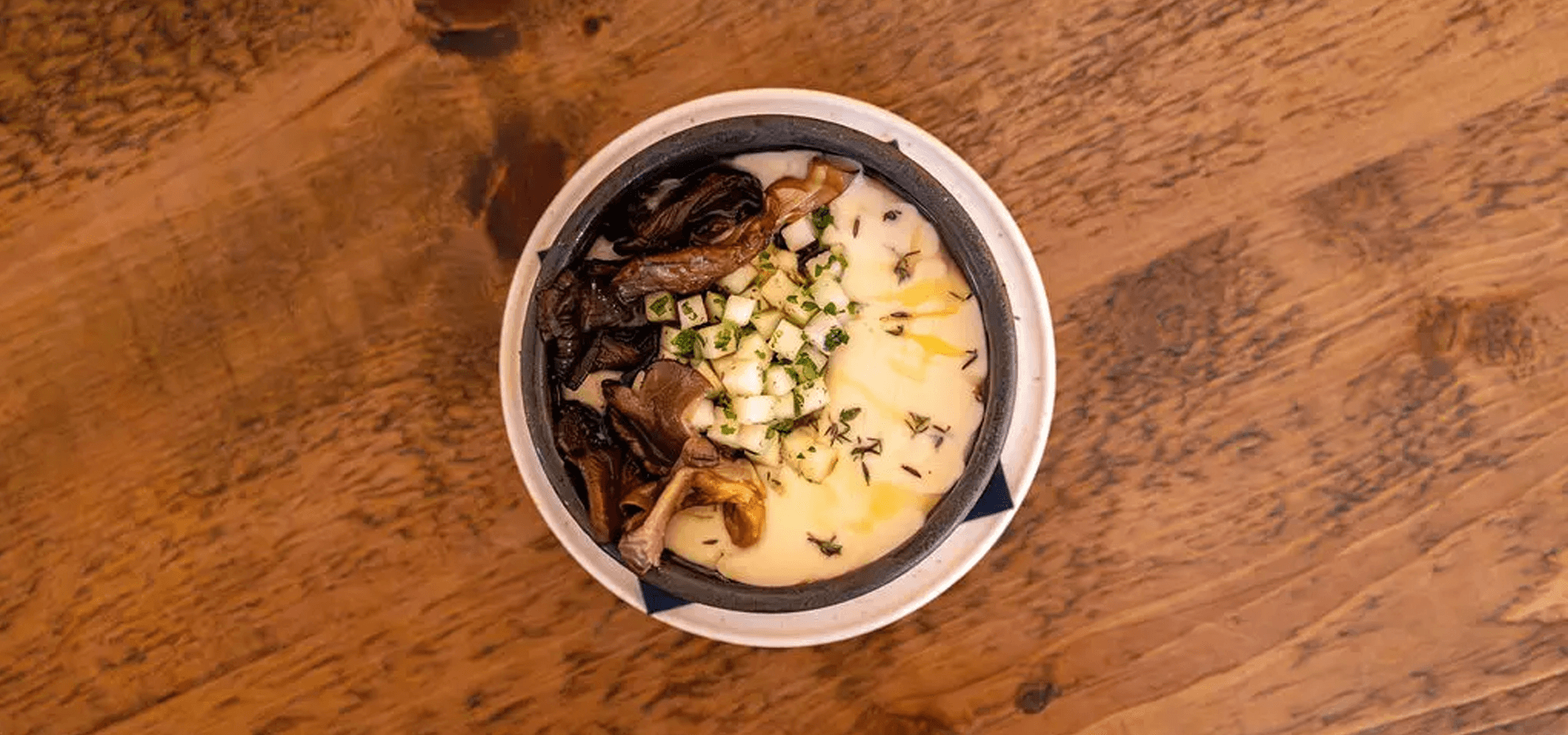 A bowl of creamy soup topped with mushrooms and herbs on a wooden table.