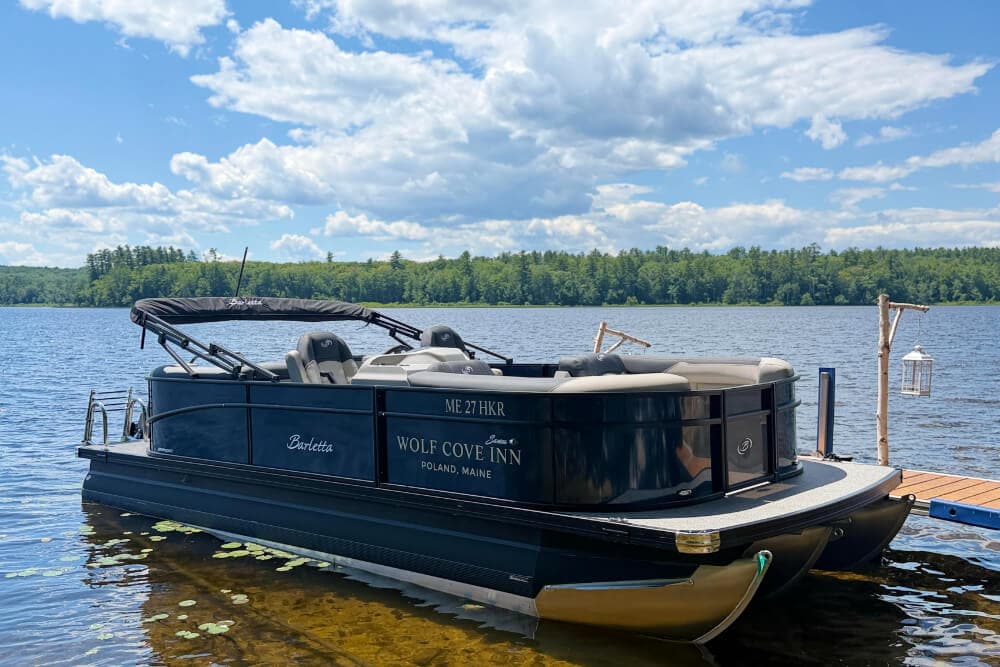 A black pontoon boat named "Barietta" docked by a serene lake under a blue sky with fluffy clouds.