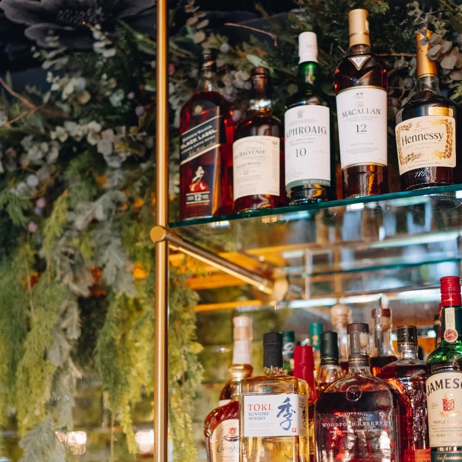 A glass shelf displays various whiskey and liquor bottles against a backdrop of greenery.