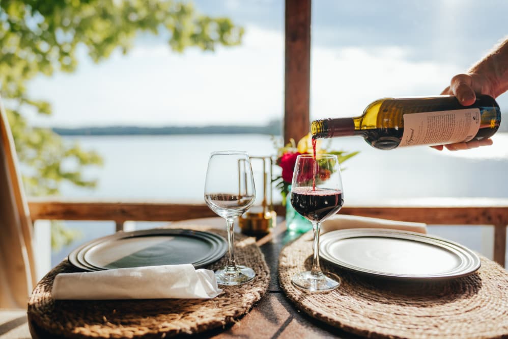 A hand pours red wine into a glass at a lakeside dining table set for two.