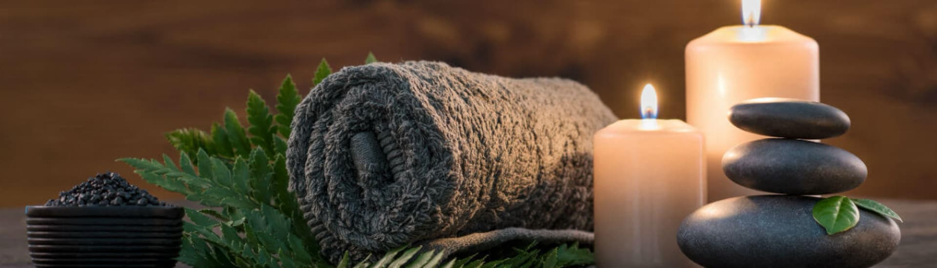 A rolled gray towel sits among candles, stacked stones, and green leaves on a wooden surface.