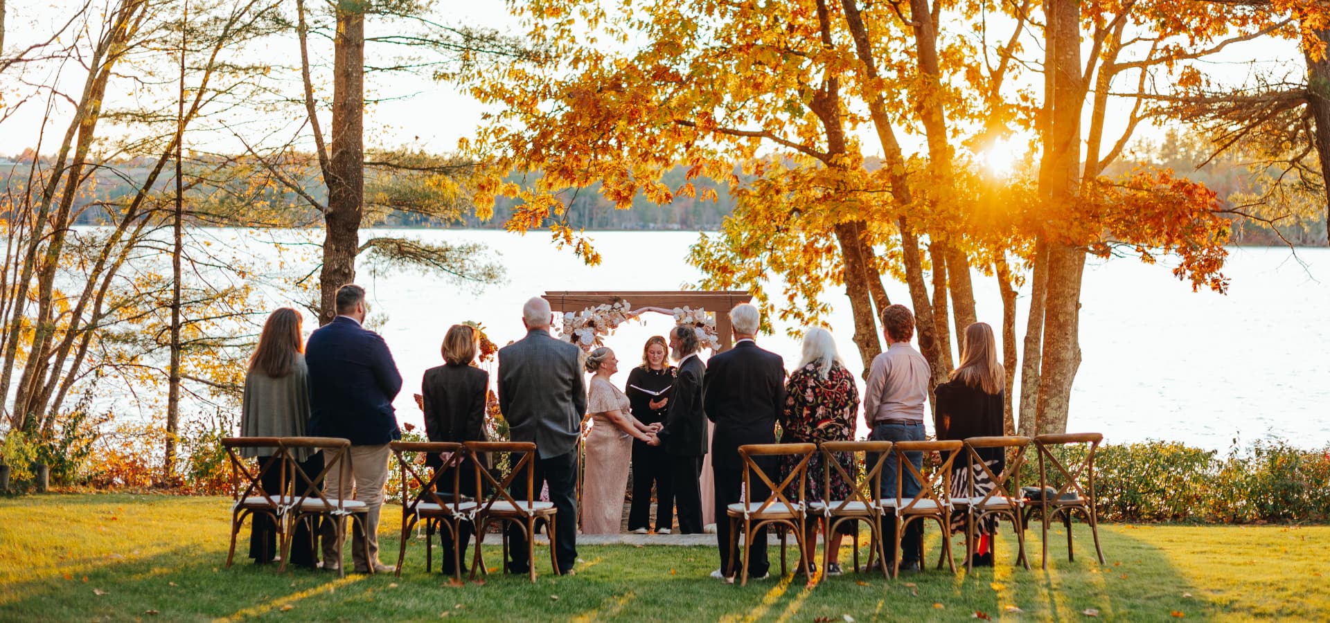 A couple stands under an arch, surrounded by guests, during a sunset wedding ceremony by the lake.