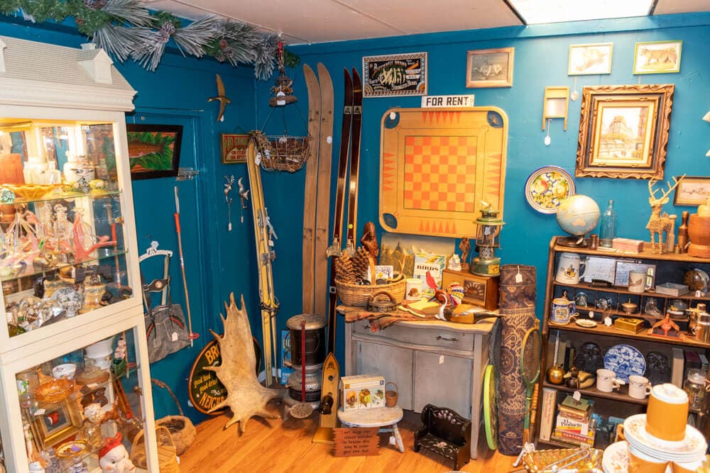 A cluttered vintage shop corner featuring decorative items, skis, a vintage checkerboard, and various antiques against a blue wall.