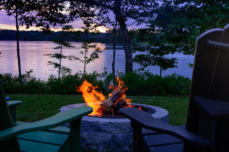 A cozy fire burns in a stone pit near a calm lake at sunset, surrounded by green chairs and trees.