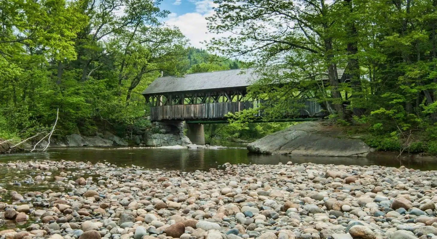 A covered bridge spans a stream surrounded by lush greenery and smooth stones.