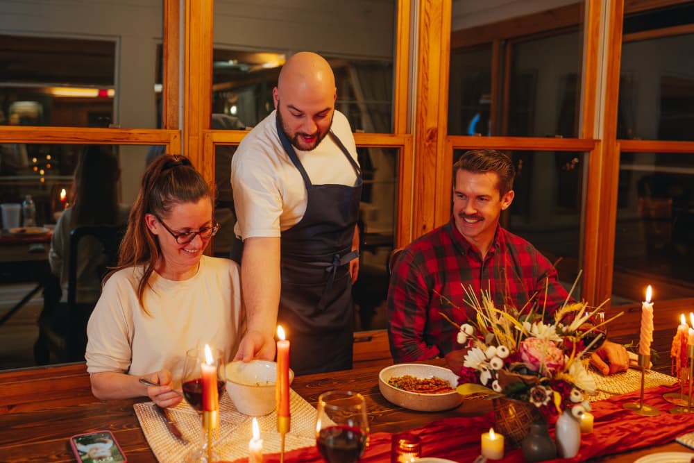 A chef serves food at a cozy dinner table surrounded by friends, with candles and a floral centerpiece.