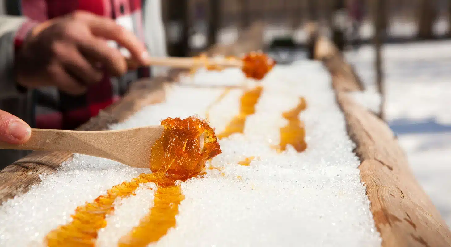 Hands using wooden sticks to drizzle syrup over a bed of snow.